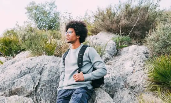 A man wearing glasses and a backpack sits on a rock, enjoying the outdoors in a serene environment, a visual representation of one of the I go crazy for hinge responses