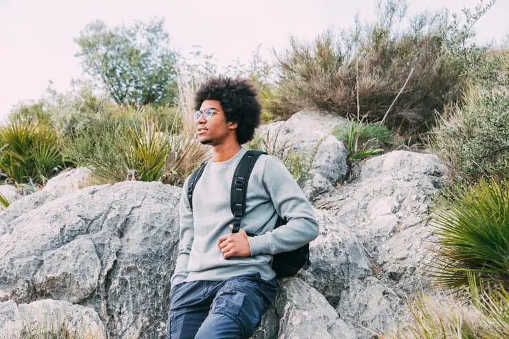 A man wearing glasses and a backpack sits on a rock, enjoying the outdoors in a serene environment, a visual representation of one of the I go crazy for hinge responses